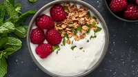 Fresh raspberries and crunchy granola top creamy yogurt bowl garnished with mint leaves on dark background, healthy breakfast or snack setup.
