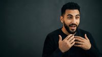 Young Middle Eastern man with dark hair and beard wearing black shirt gesturing while speaking against gray background, suitable for business communication and educational content.