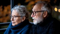 Elderly couple with gray hair and glasses sitting together in warm evening light, senior man and woman enjoying peaceful moment indoors.