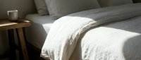 Unmade bed with white linen sheets and gray pillows bathed in natural sunlight, featuring a wooden nightstand with glass. Use for bedroom design, interior styling, hospitality, and home decor projects.