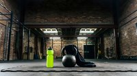 Fitness equipment arranged in industrial gym space with neon yellow water bottle, stability ball, and black resistance bands on concrete floor surrounded by brick walls and overhead lighting.