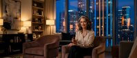 Middle-aged woman with curly blonde hair, Caucasian ethnicity, sitting in modern luxury apartment with floor-to-ceiling windows overlooking illuminated city skyline at night, holding tablet device, wearing beige blazer, relaxed professional setting with contemporary furniture and warm lighting.