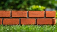 Red brick garden border wall on green grass lawn with natural bokeh background, close-up view showing masonry texture and landscaping detail.