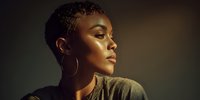Profile portrait of young African American woman with short hair and gold hoop earrings against dark background, illuminated by warm dramatic lighting highlighting facial features.