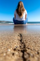 Young woman in white shirt and jeans sitting on sandy beach, view from behind with focus on sand crack pattern in foreground against blurred ocean background.