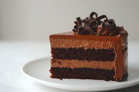 Rich chocolate layer cake slice with ganache frosting and decorative curls on white plate for bakery menu dessert photography.