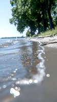Tranquil lakeshore scene with sparkling water reflections and sandy beach, trees casting shadows along shoreline as gentle waves lap against coast on sunny day.