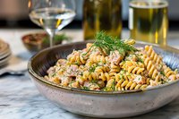 Creamy fusilli pasta with fresh shrimp, sweet corn kernels and dill garnish served in ceramic bowl on marble table, wine glass and beer in background, restaurant setting.