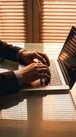 Professional businessman typing on laptop computer at modern office desk with warm sunlight streaming through venetian blinds creating shadow patterns.