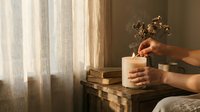 Hands lighting candle on wooden nightstand with books and dried flowers in warm bedroom interior for relaxation and cozy home atmosphere.