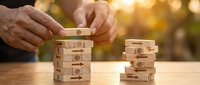 Senior businessman hands carefully placing wooden block with dollar symbol on jenga tower representing financial planning and investment strategy.