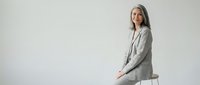 Professional mature woman with gray hair in light blazer sitting confidently on white stool against neutral background for business presentations.