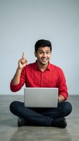 Young Indian businessman in red shirt pointing up with eureka moment while using laptop computer sitting cross-legged on floor for business presentations.