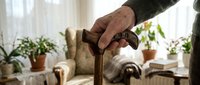 Senior man with wrinkled hands holding wooden walking cane in bright living room with potted plants and comfortable furniture, depicting aging, mobility assistance, and home care.
