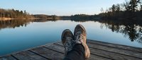 Feet in hiking shoes resting on a wooden dock overlooking a calm lake surrounded by trees under a clear blue sky.