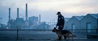 Security guard with German Shepherd dog patrolling industrial area at dusk with factory smokestacks and urban skyline in background for safety surveillance.