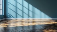 Empty room with sunlight casting geometric shadows through window on dark herringbone parquet flooring and teal wall, creating dramatic minimalist interior space.