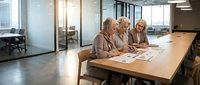 Mature women reviewing documents at wooden table in modern office with glass partitions and natural light from windows.