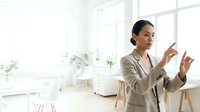 Young South Asian woman in business casual attire presenting ideas in bright, modern office space with plants and natural light for corporate training and professional development content.