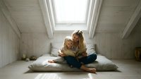 Young mother and daughter reading book together on bed in bright attic bedroom with skylight window, family bonding time and education concept.