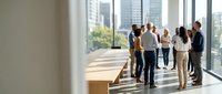 Diverse group of business professionals standing in modern office hallway with floor-to-ceiling windows overlooking cityscape, discussing corporate strategy and collaboration.