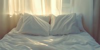 Unmade bed with white linens and pillows in morning sunlight streaming through sheer curtains, creating soft shadows and warm atmosphere for lifestyle content.