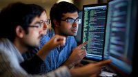 Two software developers in blue shirts working together at computer monitors displaying colorful programming code in dark office environment.