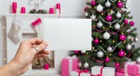 Hand holding blank white card mockup with pink and white Christmas tree decorations in cozy holiday living room interior for seasonal greeting design.