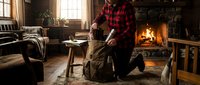 Adult male wearing red plaid shirt and child in olive jacket by fireplace in rustic cabin interior with warm lighting and cozy home decor.