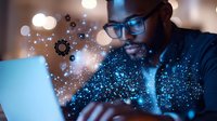 African American male professional in glasses working on laptop with glowing digital particles and gears overlay, representing technology innovation and digital transformation.
