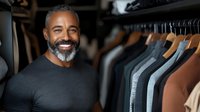 Mature African American man with gray beard smiling in clothing store, wearing black t-shirt. Professional fashion retail environment with hanging garments in background.