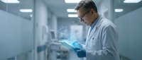Middle-aged Caucasian male scientist in white lab coat and safety glasses examining tablet computer in modern sterile laboratory environment for research work.