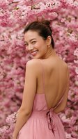 Young Asian woman in pink sundress smiling over shoulder against blooming cherry blossom background, perfect for spring fashion or beauty advertising.