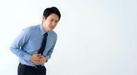 Young Asian businessman in blue shirt and tie holding stomach in pain, experiencing abdominal discomfort isolated on white background for health concepts.