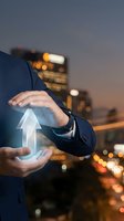 Businessman in formal blue suit holding glowing upward arrow symbol representing growth, success, and financial progress against blurred city skyline at dusk for business concepts.