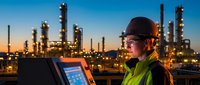 Industrial engineer in safety gear monitoring refinery operations on digital tablet during sunset with petrochemical plant towers and equipment in background.