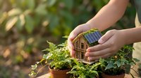 Hands holding a small wooden house model with solar panels surrounded by green plants, symbolizing eco-friendly living and sustainable energy concepts.