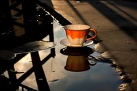 Orange and white porcelain teacup with saucer reflected in puddle water on street, dramatic shadows and sunlight creating moody atmosphere.
