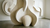 Modern abstract sculptural forms in neutral beige tones creating flowing organic shapes on concrete floor in minimalist gallery interior space.