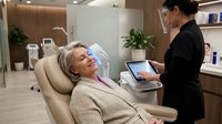 Middle-aged Caucasian woman receiving professional facial treatment from Asian female aesthetician in modern medical spa clinic with advanced equipment.