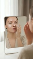 Young woman of East Asian descent applying skincare cream to her face while looking at mirror reflection, demonstrating daily beauty and wellness routine for cosmetic and dermatology marketing.