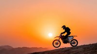 Motocross rider jumping dirt bike silhouette against vibrant orange sunset sky over mountain landscape for extreme sports and adventure content.