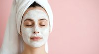 Young woman with white clay facial mask and towel on head relaxing with closed eyes against pink background for skincare routine and spa treatments.