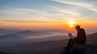 Young man working on laptop computer while sitting on mountain peak during golden hour sunrise with layered hills and misty valleys below.