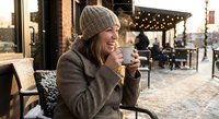 Young woman enjoying hot beverage at outdoor cafe terrace during winter evening with warm lighting and cozy atmosphere for lifestyle content.