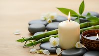 Spa wellness composition with burning candle, smooth stones, bamboo stems, jasmine flowers and wooden bowl on natural background for relaxation.