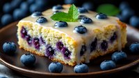 Fresh blueberry cake slice with vanilla glaze and mint garnish on brown plate, surrounded by ripe berries, close up view showing moist texture and fruit filling.