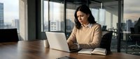 Young South Asian woman working at wooden desk with laptop and notebook in modern high-rise office with city skyline view, professional business environment.