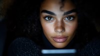 Young African American woman with natural curly hair and freckles illuminated by soft blue light from digital device screen in dark environment.
