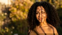 Young African American woman with curly hair smiling in warm golden sunlight during autumn season outdoors with natural bokeh background.